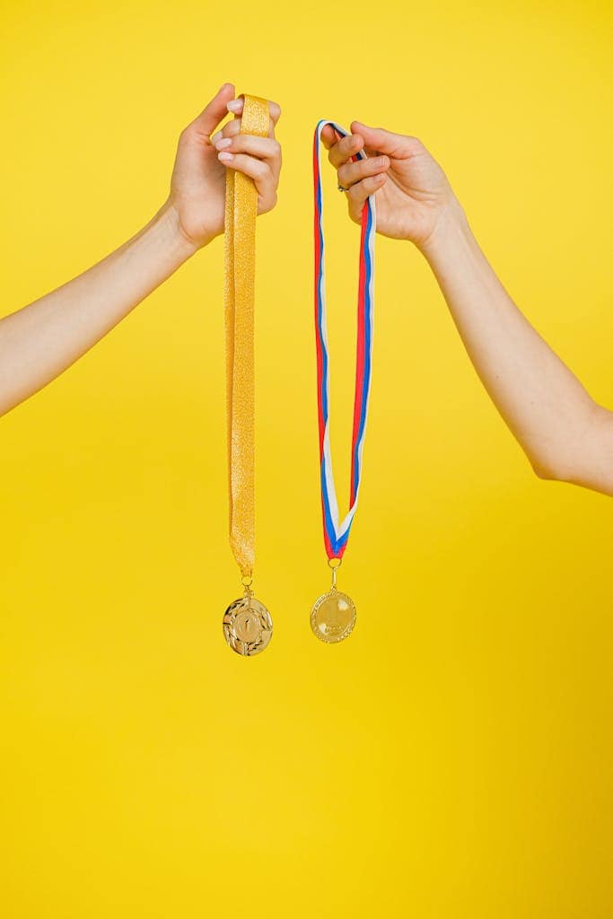 Two hands holding gold and silver medals on a vibrant yellow background, symbolizing success and achievement.