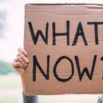 Cardboard sign reading 'What Now?' held outdoors, conveying uncertainty or protest.