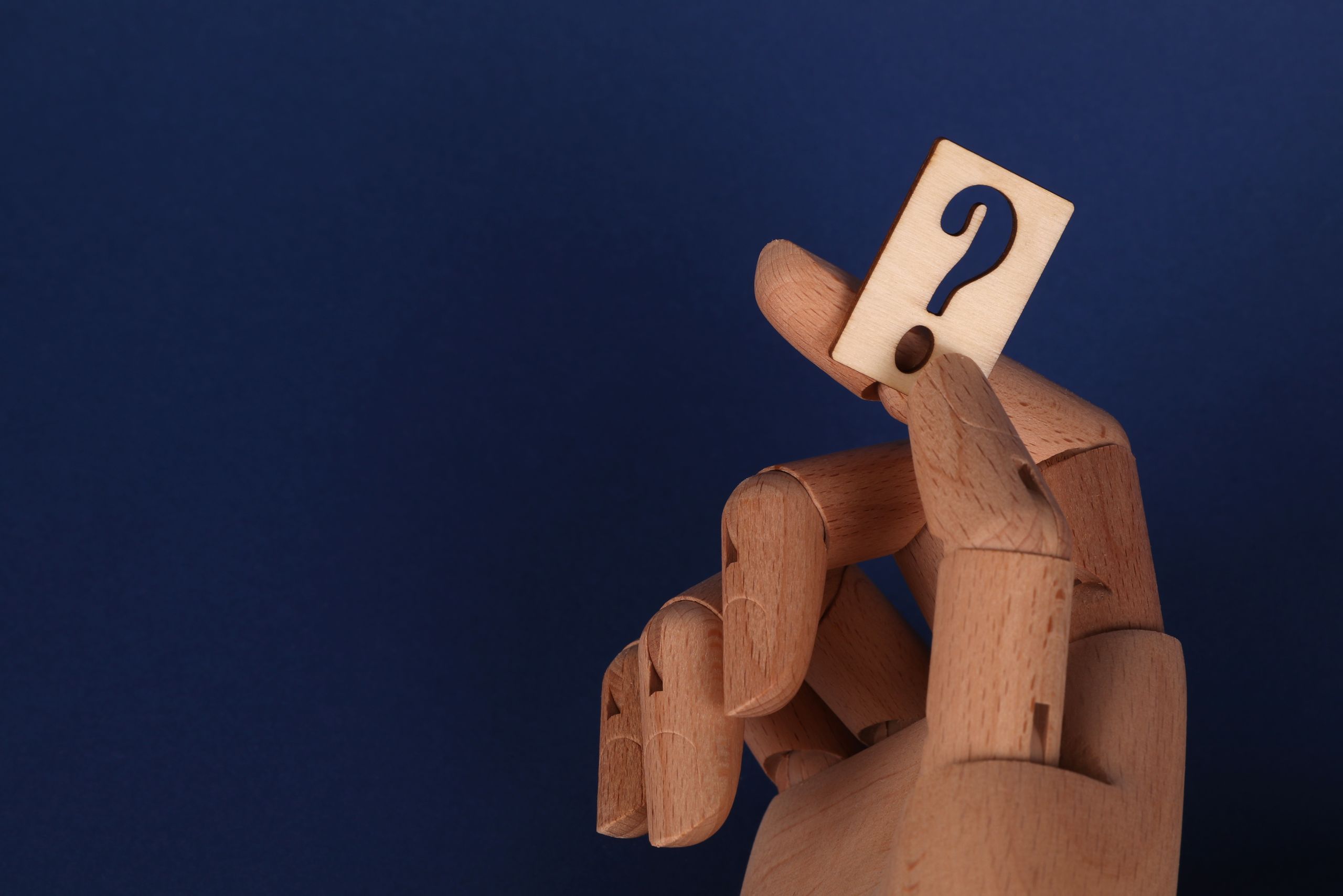 Wooden mannequin hand holding a block with a question mark against a blue background.