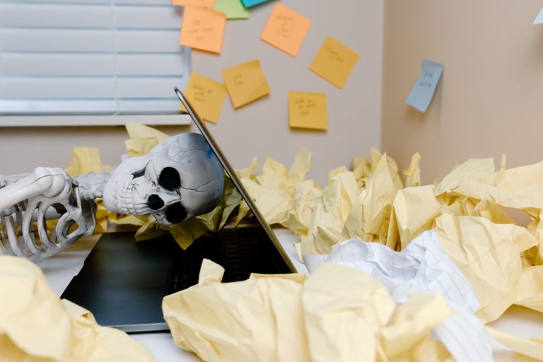 A humorous skeleton slumped over a laptop among crumpled papers in an office setting.