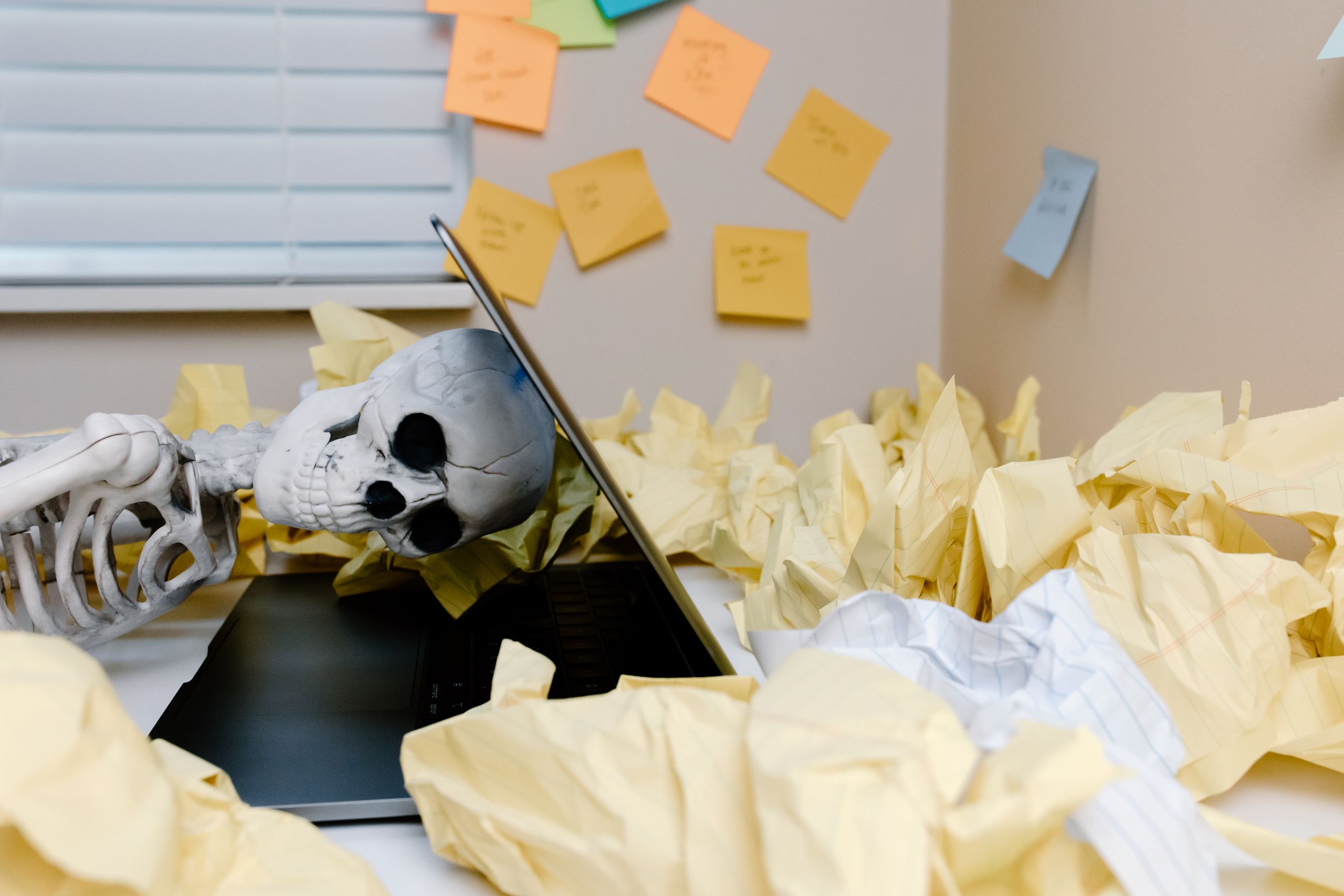 A humorous skeleton slumped over a laptop among crumpled papers in an office setting.