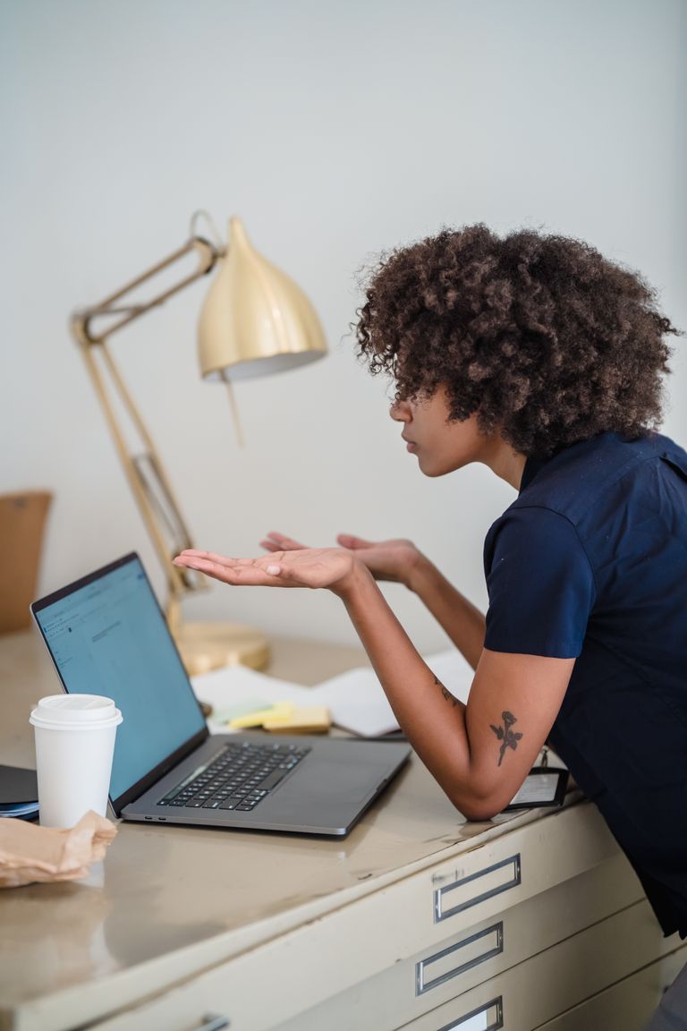 Woman sitting at desk with laptop, gesturing in confusion, inside office setting.