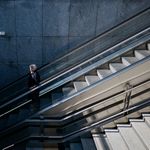 A lone person ascends an urban escalator amidst sleek, modern architecture.