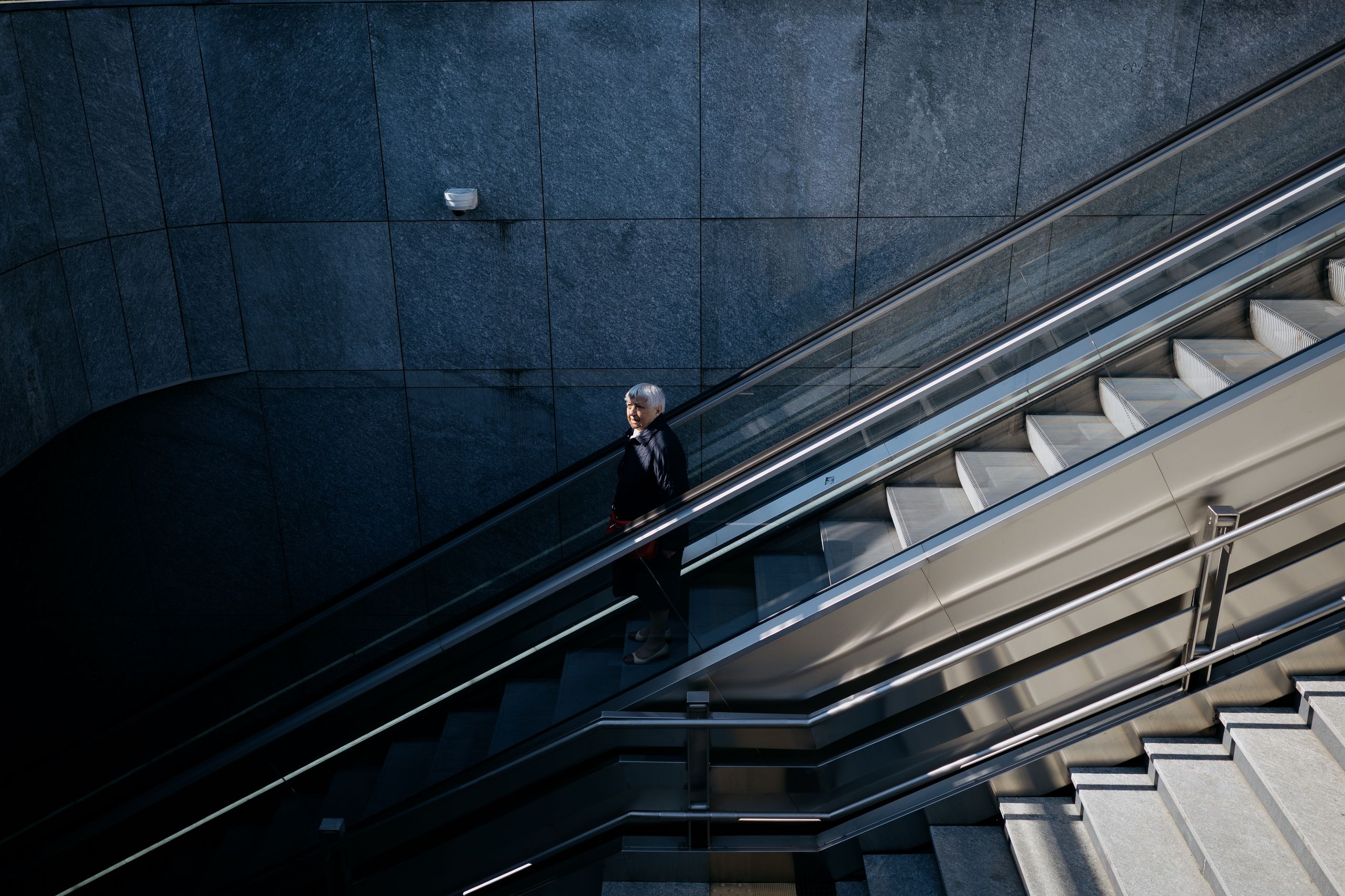 A lone person ascends an urban escalator amidst sleek, modern architecture.