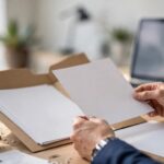 A professional compares blank tender documents beside a laptop and green glass paperweight.